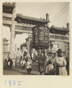 Members of a funeral procession carrying an umbrella and paper snow willows under a pai lou