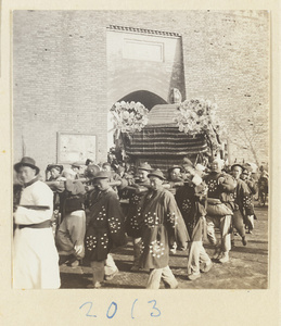 Coffin-bearers carrying draped coffin with wreaths of paper flowers through city gate during funeral procession