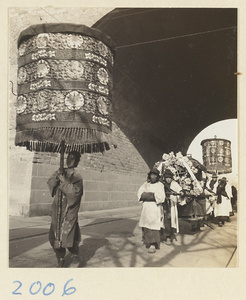 Members of a funeral procession carrying umbrellas and litter through archway of a city gate