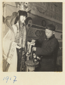 Boy placing basket of flowers in the hands of a paper figure for a funeral procession