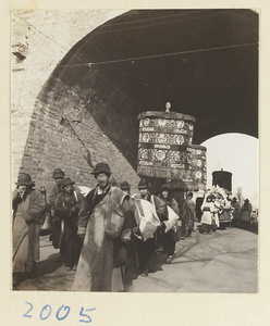 Members of a funeral procession carrying umbrellas and litter through archway of a city gate