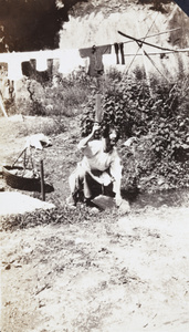 Women using a paddle to clean laundry in a stream, near a bamboo drying rack