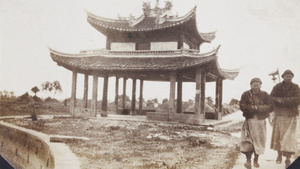 Two monks walking past a pavilion and grave mounds