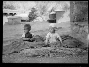 Yinhua, the baby son of Guo Qinglan and Dr Dwarkanath S. Kotnis, sitting on a  sheet on top of some hay, with another baby, Jinchaji